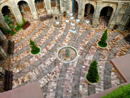 A beautifully patterned stone courtyard with concentric circular designs. The space features several green coniferous trees planted in circular patches of grass. A central circular stone fountain is visible, surrounded by decorative hanging lights. Arched entrances made of stone line the perimeter of the courtyard.