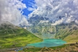 A tranquil turquoise lake is nestled among lush green hills. The backdrop consists of dramatic, snow-capped mountains partially shrouded in clouds under a vibrant blue sky. Small tents are scattered along the lakeside, indicating a campsite area.