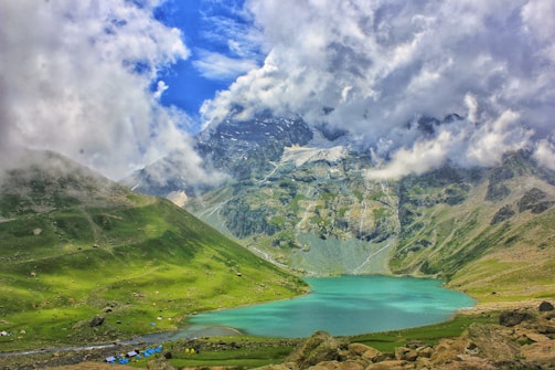 A tranquil turquoise lake is nestled among lush green hills. The backdrop consists of dramatic, snow-capped mountains partially shrouded in clouds under a vibrant blue sky. Small tents are scattered along the lakeside, indicating a campsite area.