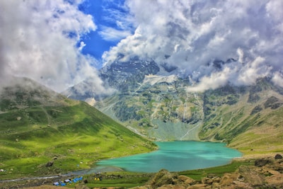 A tranquil turquoise lake is nestled among lush green hills. The backdrop consists of dramatic, snow-capped mountains partially shrouded in clouds under a vibrant blue sky. Small tents are scattered along the lakeside, indicating a campsite area.
