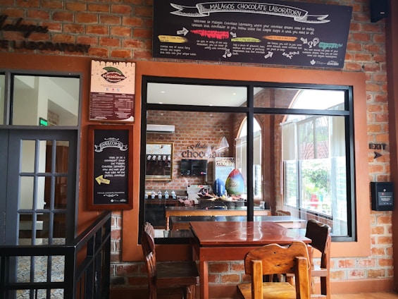 An inviting interior of a chocolate laboratory featuring a wooden table and chairs in the foreground. The room has brick walls adorned with various signs and informational boards, prominently displaying the name 'Malagos Chocolate Laboratory.' Large windows provide a view into the chocolate preparation area, which includes decorative chocolate eggs and other chocolate-making elements. The laboratory has a cozy, rustic atmosphere, emphasized by the warm color palette and wooden elements.