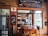 An inviting interior of a chocolate laboratory featuring a wooden table and chairs in the foreground. The room has brick walls adorned with various signs and informational boards, prominently displaying the name 'Malagos Chocolate Laboratory.' Large windows provide a view into the chocolate preparation area, which includes decorative chocolate eggs and other chocolate-making elements. The laboratory has a cozy, rustic atmosphere, emphasized by the warm color palette and wooden elements.