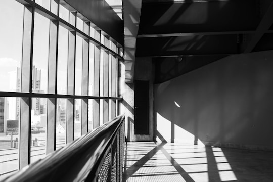 A black and white interior space features large windows on the left side, allowing ample natural light to cast geometric shadows on the floor and walls. The view through the windows reveals an urban skyline, including several high-rise buildings. The architecture is modern with clean lines and a minimalist aesthetic.