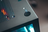 A close-up view of a computer tower, featuring a power button and ventilation holes on the top. The LED lights emit a blue and red glow, adding a modern and high-tech feel. The angled perspective highlights the sleek, metallic surface of the case.