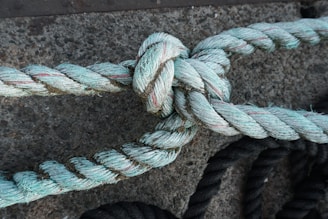 A close-up view of a thick, weathered rope tied in a knot against a rough, textured concrete surface. The rope is predominantly light turquoise with faint red strands, suggesting wear and exposure to the elements.