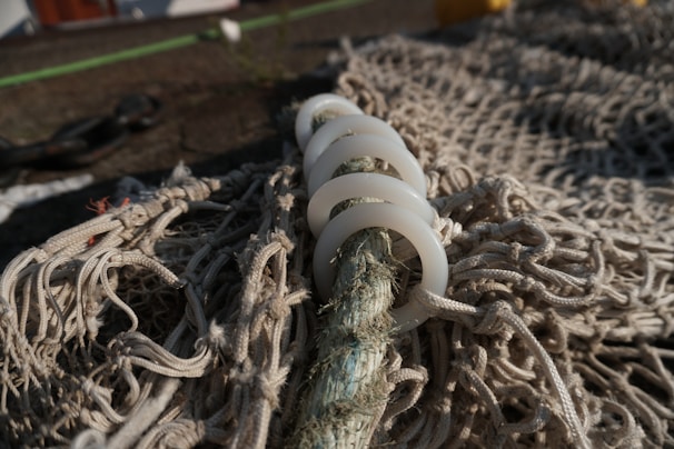 A detailed view of a fishing net with white plastic rings threaded onto a rope. The net is spread out on a surface with blurred background elements, such as other ropes and indistinct objects.