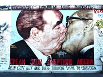 A mural depicting two men engaging in a kiss, with various graffiti and messages written across their faces and around the image. The text below is in multiple languages, including Russian and German. The background is filled with colorful graffiti.