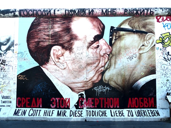 A mural depicting two men engaging in a kiss, with various graffiti and messages written across their faces and around the image. The text below is in multiple languages, including Russian and German. The background is filled with colorful graffiti.