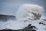 A massive wave crashes against a seawall near a lighthouse. The power of the ocean is evident as water splashes high into the air, creating a dramatic scene. The sky is overcast, contributing to the turbulent atmosphere.
