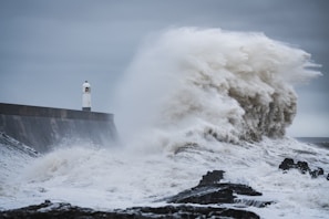 A massive wave crashes against a seawall near a lighthouse. The power of the ocean is evident as water splashes high into the air, creating a dramatic scene. The sky is overcast, contributing to the turbulent atmosphere.