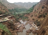 A lush river valley surrounded by rocky cliffs and greenery. Numerous small huts and umbrellas line the riverside, and people can be seen swimming and relaxing along the water. The landscape features earthy tones with rugged terrain and dense foliage.