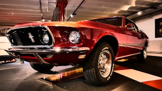 A polished red classic car is parked inside a showroom with distinct chrome accents and round headlights. The car's tires display the branding 'COBRA GT Radial'. The setting is a well-lit indoor area with a reflective floor and overhead lights.