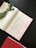 An open notebook with a red border is placed on a dark surface. On the left page, green leaves are arranged neatly. The right page features a minimalist line drawing of a face and hands in red ink, accompanied by handwritten red text.
