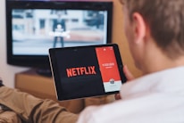 A person is sitting on a couch holding a tablet that displays the Netflix logo prominently. In the background, a television is showing a blurred image. The person appears to be engaged in using both devices simultaneously.