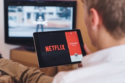 A person is sitting on a couch holding a tablet that displays the Netflix logo prominently. In the background, a television is showing a blurred image. The person appears to be engaged in using both devices simultaneously.
