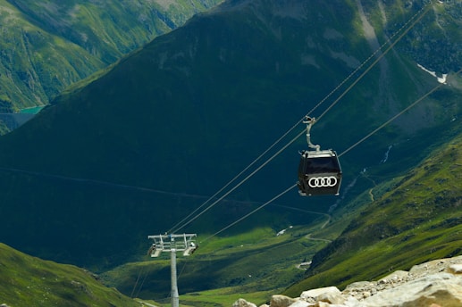 A cable car with the Audi logo travels along a cable suspended over a lush green mountainous landscape. The scene is filled with steep slopes and valleys, and there is a metal support tower in the middle distance.