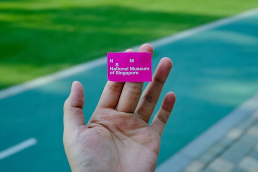A hand is holding a small pink ticket with the text 'National Museum of Singapore' visible. The background consists of a blurred outdoor pathway and green grassy area.