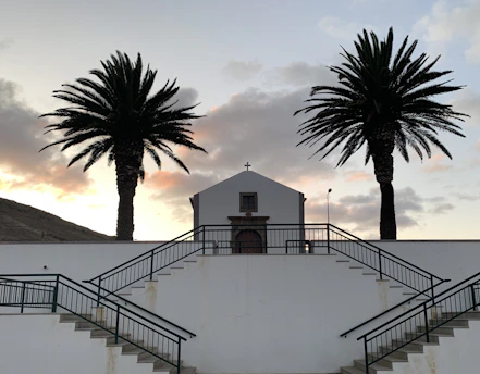 A small church is centered between two tall palm trees. The building is white with a simple wooden door, positioned on a platform accessed by concrete stairs with dark railings. The sky is a soft gradient of evening light with scattered clouds.
