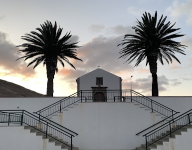 A small church is centered between two tall palm trees. The building is white with a simple wooden door, positioned on a platform accessed by concrete stairs with dark railings. The sky is a soft gradient of evening light with scattered clouds.