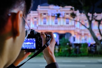 A person is holding a camera and taking a photograph of a beautifully illuminated building at night. The camera display shows the image being captured. The background features a colorful facade with intricate architectural details emphasized by the lighting.