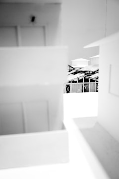 A monochromatic image featuring a model of architectural structures. The focus appears to be on angular buildings with visible geometric patterns and rooftop details. The blurred foreground creates a sense of depth and frames the central elements.