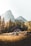 A rustic wooden cabin is nestled in a serene mountainous landscape, surrounded by lush green pine trees. In the background, towering rocky mountains rise under a bright sky. The foreground features a grassy meadow with a few large rocks scattered around.