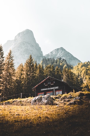 A warm, rustic cabin nestled among pine trees with a backdrop of towering alpine peaks at sunset.