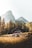 A rustic wooden cabin is nestled in a serene mountainous landscape, surrounded by lush green pine trees. In the background, towering rocky mountains rise under a bright sky. The foreground features a grassy meadow with a few large rocks scattered around.