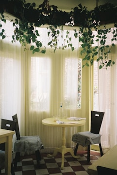 A dining area featuring a muted yellow tablecloth, eclectic chairs, and potted plants on shelves.