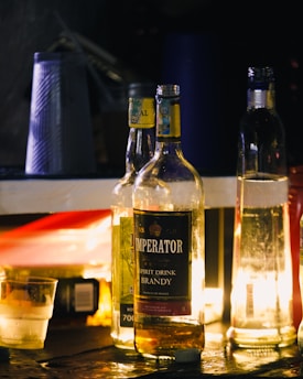 A collection of alcohol bottles, including one labeled 'Brandy,' are arranged on a surface with multiple lighting sources, creating a warm and inviting glow. Nearby are some plastic cups with a textured pattern, and a small glass with ice is also visible.