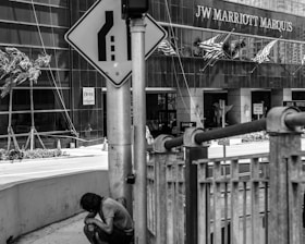A person is sitting on the sidewalk beside a metal railing and a traffic sign near a large hotel building. The hotel has American flags displayed above the entrance and is identified as the JW Marriott Marquis. Palm trees and various signs are visible nearby.