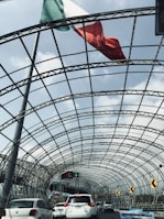 A large Mexican flag waves above a metal mesh tunnel structure over a busy road with several cars in traffic. The sky is partly cloudy, and there are road signs and streetlights visible.