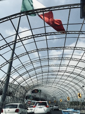 A large Mexican flag waves above a metal mesh tunnel structure over a busy road with several cars in traffic. The sky is partly cloudy, and there are road signs and streetlights visible.
