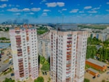 High-rise residential buildings with multiple stories, surrounded by other apartment complexes, greenery, and parked cars. The background displays a vast landscape with more buildings, trees, and a distant smoky horizon under a blue sky dotted with clouds.
