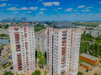 High-rise residential buildings with multiple stories, surrounded by other apartment complexes, greenery, and parked cars. The background displays a vast landscape with more buildings, trees, and a distant smoky horizon under a blue sky dotted with clouds.