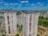 High-rise residential buildings with multiple stories, surrounded by other apartment complexes, greenery, and parked cars. The background displays a vast landscape with more buildings, trees, and a distant smoky horizon under a blue sky dotted with clouds.