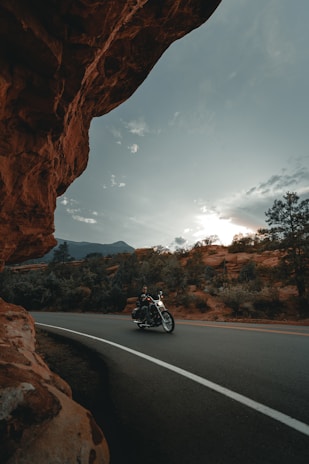 A motorcyclist rides along a curved road surrounded by red rock formations and dense vegetation. The sky above is partly cloudy with the sun peeking through, casting a warm glow over the landscape.