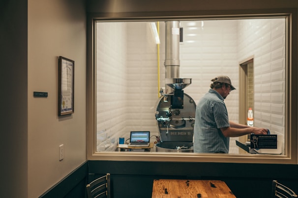 A person wearing a cap is operating a coffee roasting machine in a small, tidy room with tiled walls. A laptop is visible on a small table beside the machine, and a sign on the wall indicates occupancy details. The room has a clean and modern appearance, with a framed document on the outside wall.