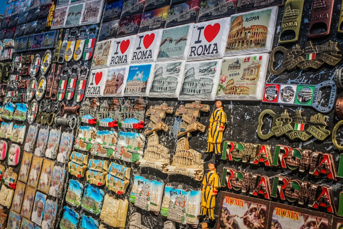 A collection of colorful souvenirs featuring various themes related to Rome. There are magnets, keychains, and plaques with the Colosseum, Vatican, and inscriptions like 'I Love Roma' and 'Italia'. Brightly colored Italian flags and Roman numerals are also prominent.