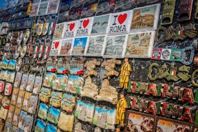 A collection of colorful souvenirs featuring various themes related to Rome. There are magnets, keychains, and plaques with the Colosseum, Vatican, and inscriptions like 'I Love Roma' and 'Italia'. Brightly colored Italian flags and Roman numerals are also prominent.
