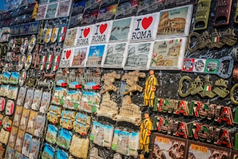 A collection of colorful souvenirs featuring various themes related to Rome. There are magnets, keychains, and plaques with the Colosseum, Vatican, and inscriptions like 'I Love Roma' and 'Italia'. Brightly colored Italian flags and Roman numerals are also prominent.