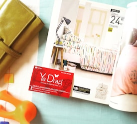 A neatly arranged scene featuring a magazine open to a page showcasing a colorful bedspread in a modern bedroom setting. Beside the magazine, there is a green leather wallet and an orange household object. A red business card is placed on the page, providing contact information for a company called Ya Drap.