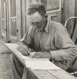 An elderly man writing thoughtfully at a wooden desk surrounded by books.