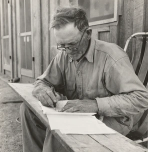 An elderly man writing thoughtfully at a wooden desk surrounded by books.