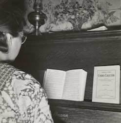 A person with glasses stands in front of a piano, reading a music sheet. The piano has a decorative floral backdrop and a small vase with flowers on top. There is a sheet music booklet labeled 'Chorus Collection' placed on the piano alongside the open sheet music.