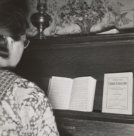 A person with glasses stands in front of a piano, reading a music sheet. The piano has a decorative floral backdrop and a small vase with flowers on top. There is a sheet music booklet labeled 'Chorus Collection' placed on the piano alongside the open sheet music.