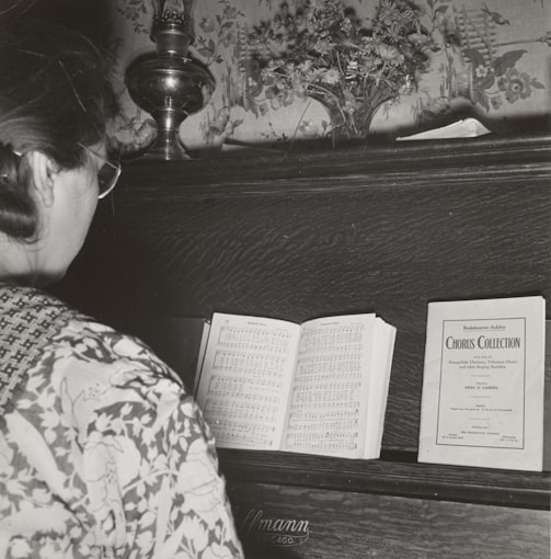 A person with glasses stands in front of a piano, reading a music sheet. The piano has a decorative floral backdrop and a small vase with flowers on top. There is a sheet music booklet labeled 'Chorus Collection' placed on the piano alongside the open sheet music.