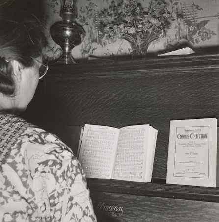 A person with glasses stands in front of a piano, reading a music sheet. The piano has a decorative floral backdrop and a small vase with flowers on top. There is a sheet music booklet labeled 'Chorus Collection' placed on the piano alongside the open sheet music.