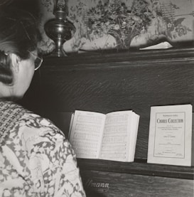 A person with glasses stands in front of a piano, reading a music sheet. The piano has a decorative floral backdrop and a small vase with flowers on top. There is a sheet music booklet labeled 'Chorus Collection' placed on the piano alongside the open sheet music.