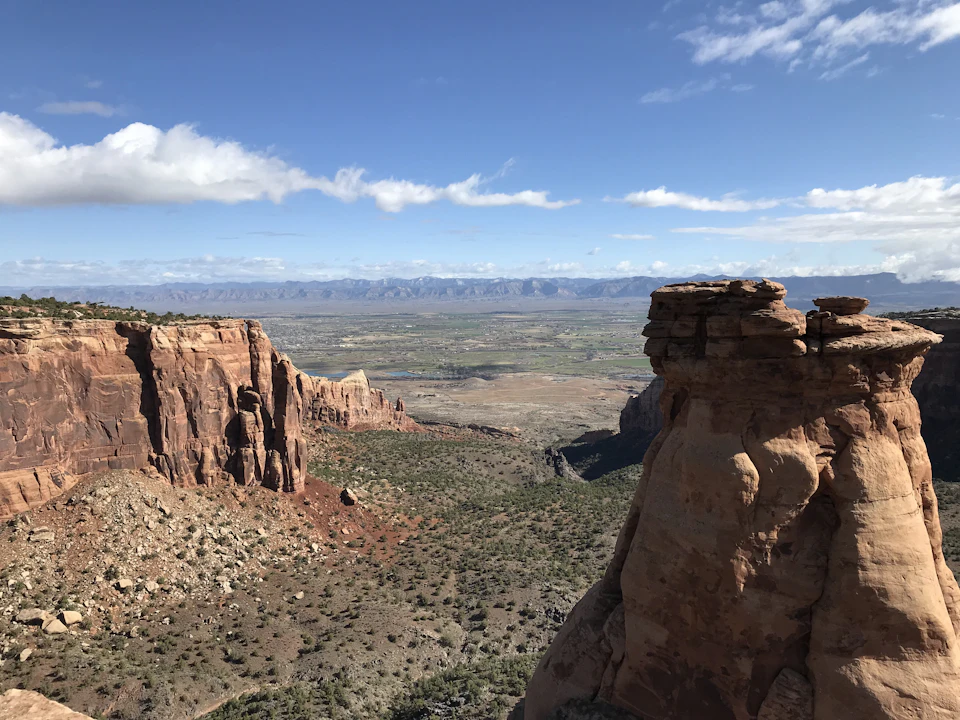 The image depicts a breathtaking view from a high vantage point, overlooking a vast desert landscape. The scene is dominated by towering red rock formations that stretch across the frame, with some cliffs appearing more rugged than others. In the distance, a valley can be seen, adding depth to the image and creating a sense of scale. The sky above is a clear blue, dotted with fluffy white clouds, providing a striking contrast against the earthy tones of the rocks below.